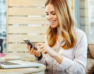 Lächelnde Frau benutzt ihr Smartphone und sitzt an einem Tisch mit einem Laptop in einem modernen Café